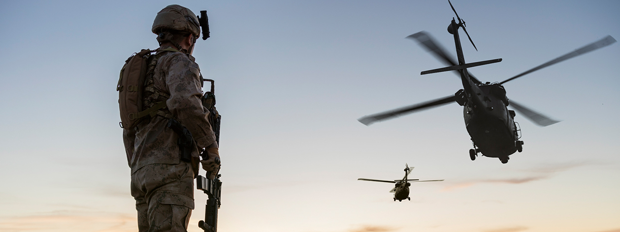Man standing in combat uniform watching 2 helicopters fly towards the sunrise