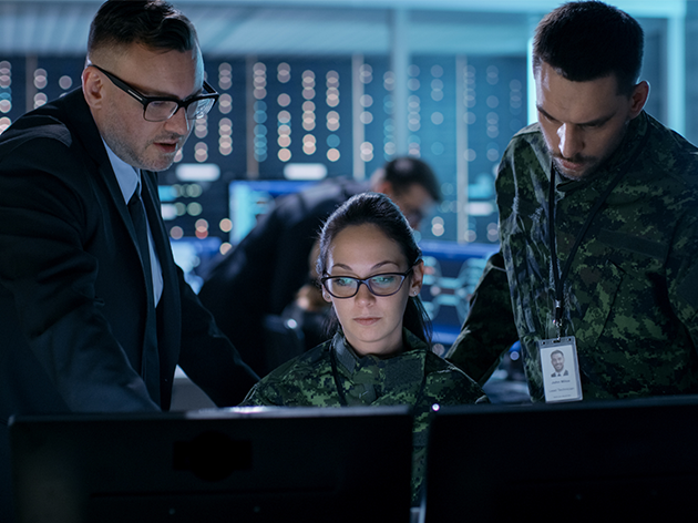 Three national security professionals confer in front of a series of computer screens in a command center