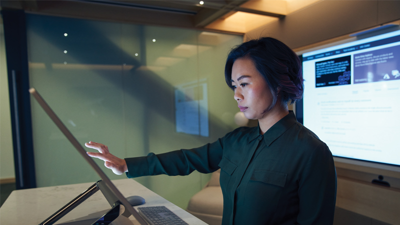 Side profile of a woman wearing a dark shirt in a dim office scrolling or working on a Microsoft Surface Studio