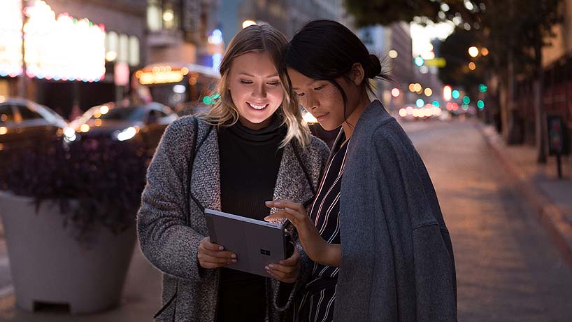 Two women on a city street interact with a Surface Go
