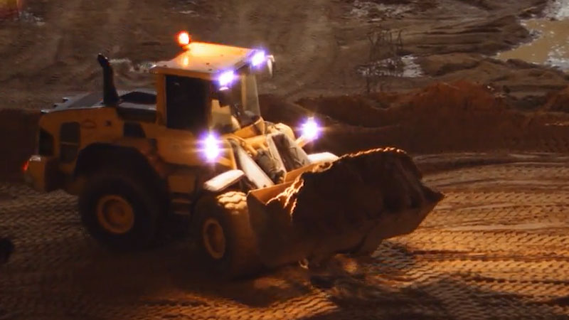 A skid-steer loader moves piles of dirt from the side of a large mound at dusk
