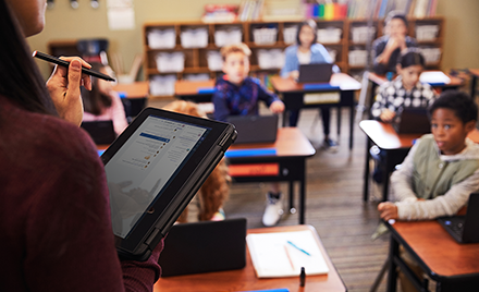 Image of a teacher checking her tablet in front of a classroom full of children at desks.