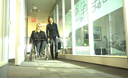 Image of two workers chatting in an accessible workplace hallway, one standing, the other from a wheelchair.