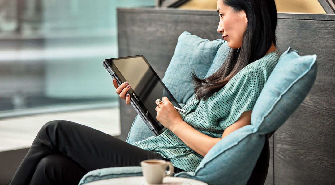 Eine Frau sitzt bequem in einer modernen Büroumgebung mit einem Kaffee und schreibt auf einem Laptop im Tablet Modus.