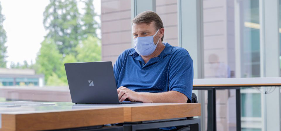 A man wearing a face mask works at a desk in front of a window in an office