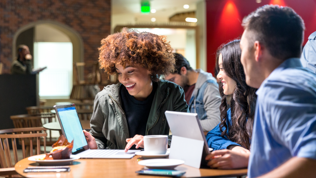 One male and two female college students sitting at round table in study café, collaborating on team project. One of the female students is smiling and using a laptop (screen blurred), while the other two look on. An open Surface Pro (screen not shown)