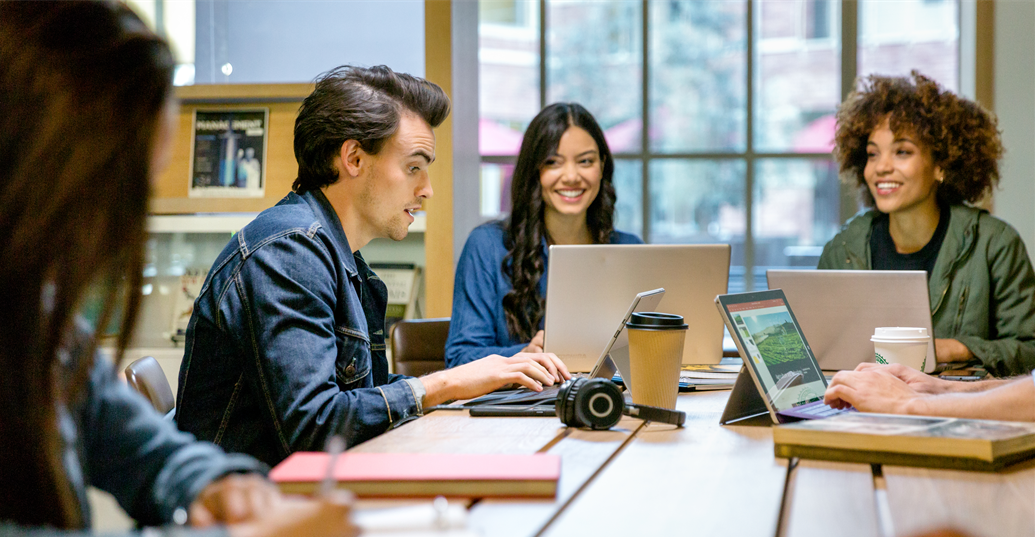 Male and female college students collaborating in group study session in library. Two Surface Pros and two laptops are on table (one Surface Pro screen shows PowerPoint slide; no other screens shown). 