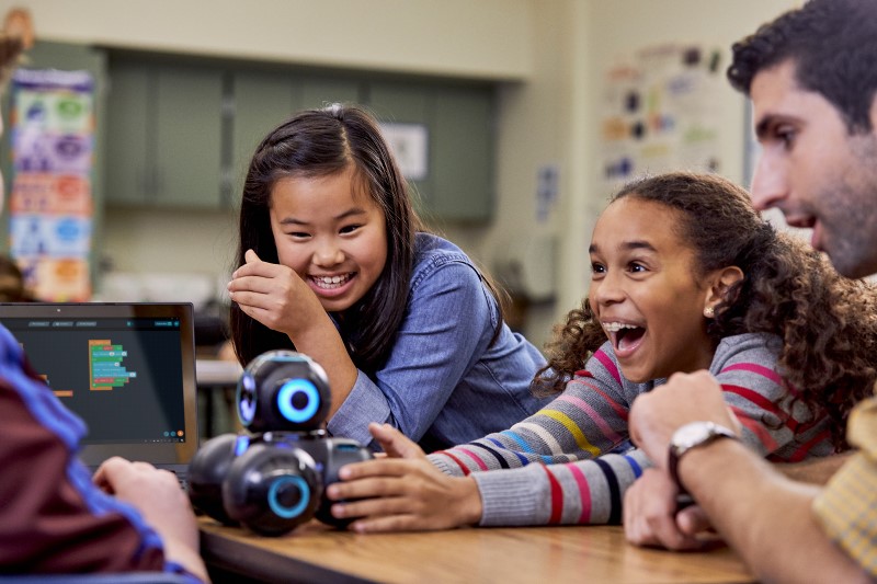 Two female students in a group including their teacher, using a Wonder Workshop Cue robot.