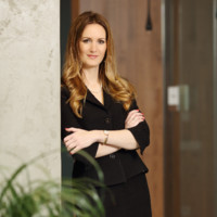 Picture of a smiling woman in a business suit, leaning against the wall with her arms folded. She has long ombre hair.