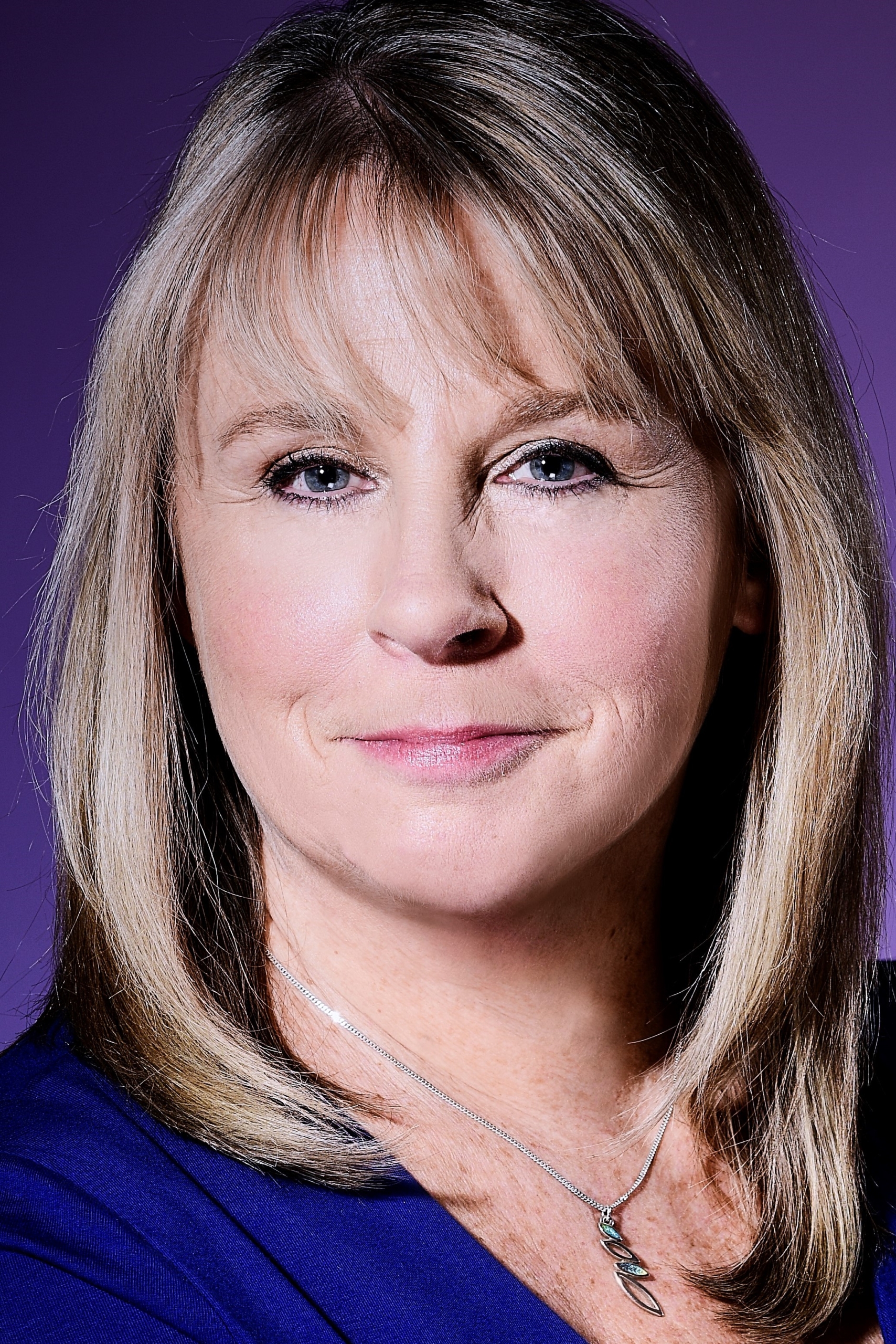 Debbie Foster, a woman wearing a blue shirt and silver necklace with dark blonde shoulder-length hair and a fringe looks at the camera and smiles. The background is dark purple.