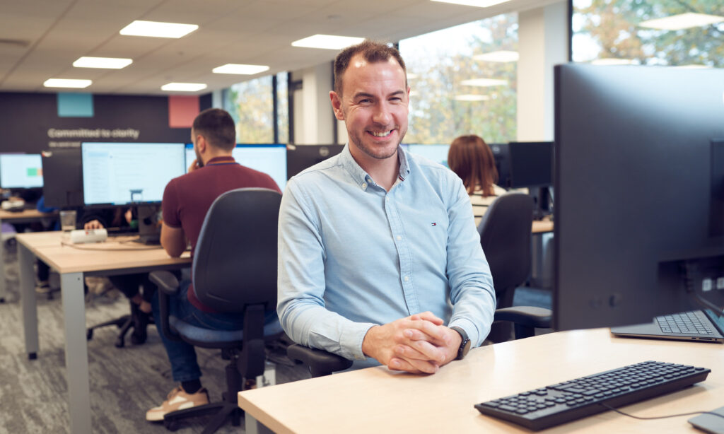 A smiling male office worker wearing a blue open-necked shirt sits at a desk