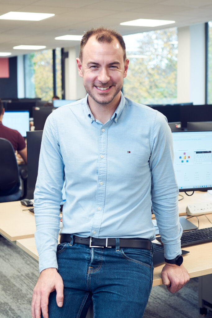 A male office worker wearing a blue open-necked shirt smiles at the camera