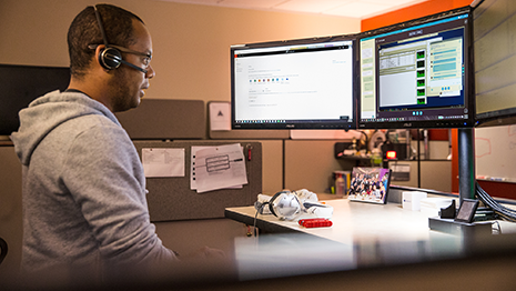 Image of a firstline worker speaking to a colleague over a headset while looking at several computer monitors.