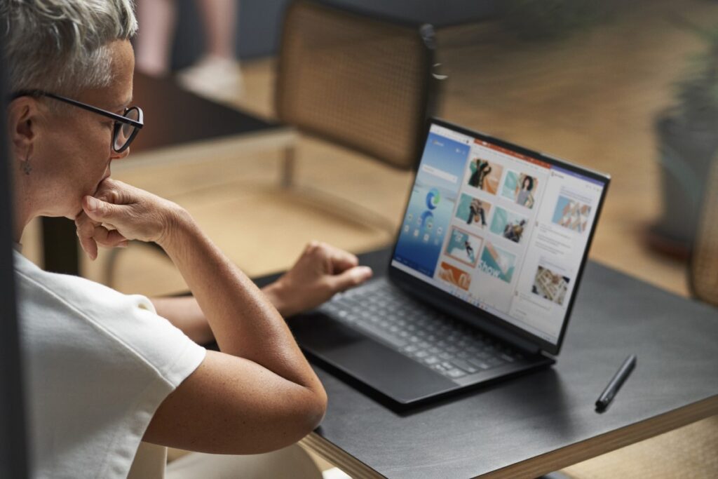 Adult sitting at table, looking at laptop with Edge, PowerPoint, and Word on screen via Snap feature. Keywords: table; over the shoulder; remote workplace; meeting; café; New York City