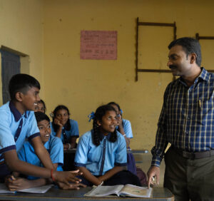 A male teacher in a blue plaid shirt interacting with students in blue uniforms in a classroom  
