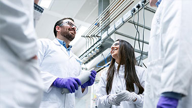 Four scientists wearing laboratory coats, purple gloves, and goggles smiling and talking together.