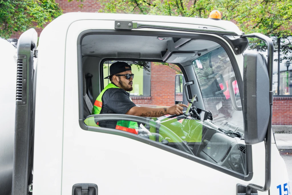 Male worker sitting in driver‘s seat of small loading truck using tablet mounted on dashboard. Photo taken from outside passenger door. Man‘s tattoos visible on arm.