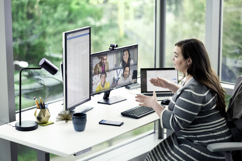 Female enterprise employee working at desk with multiple devices, including HP Elitebook, running a Microsoft Teams conference call. Two different screen images are available: one featuring 4 people on screen, another with 9 people on screen.