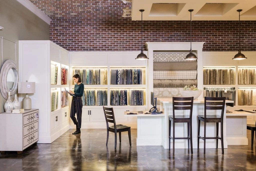 Female first line worker in retail store, standing directly in front of merchandising display wall filled with fabric panels while using pen on ASUS convertible laptop folded open as tablet (screen not shown).