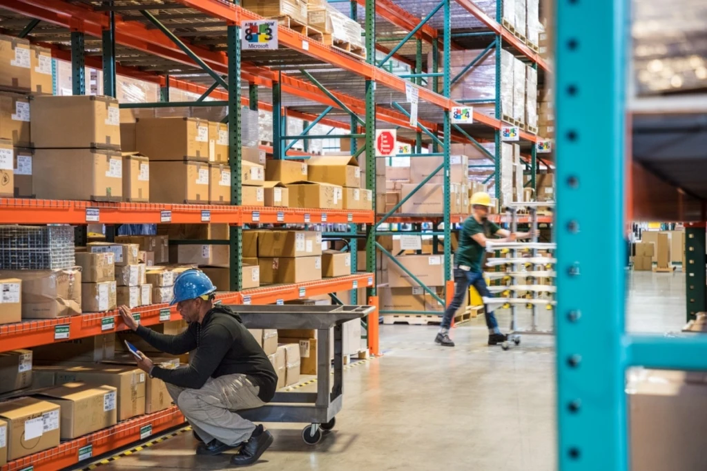 Male worker wearing hardhat using tablet in warehouse. Another worker in background.