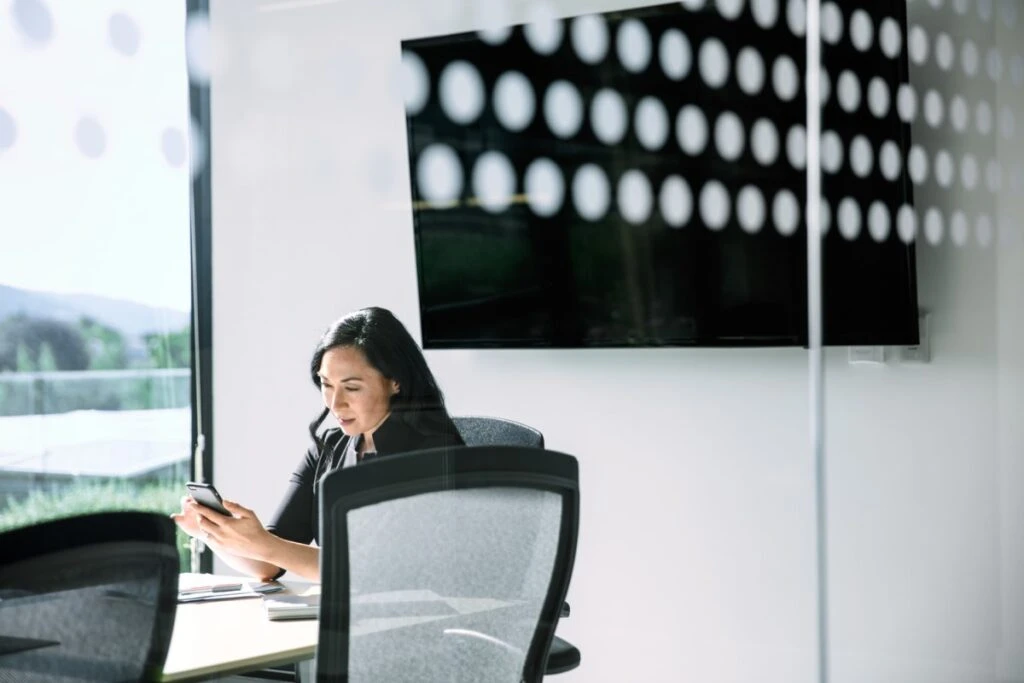 Female corporate office worker using phone at conference room table. There is a large monitor behind her (blank screen shown).