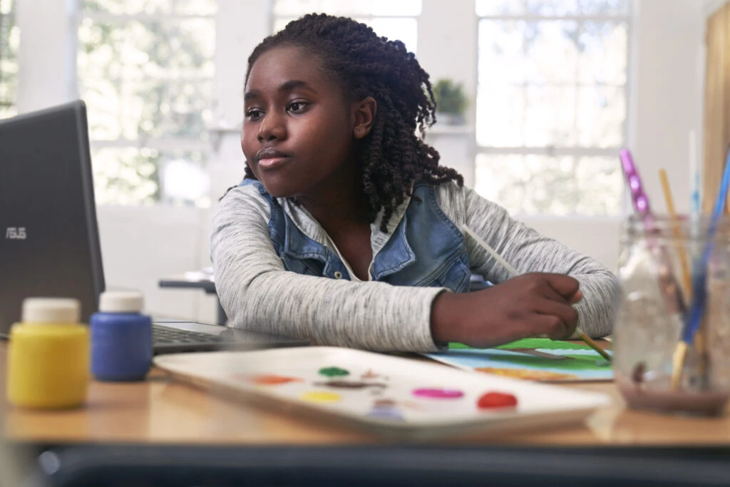 Female K-12 student sitting at a desk painting a tree while looking at a laptop.