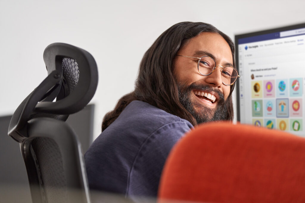 Man smiling while at work​ Keywords: Viva; Filip; hybrid work; work from home; remote; male; desktop monitor; UI screen; glasses; looking back over his shoulder; office; ; Insights; praise