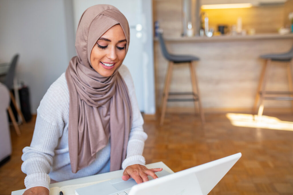 young woman sitting at a wooden table holding pencil writing on notebook next to computer