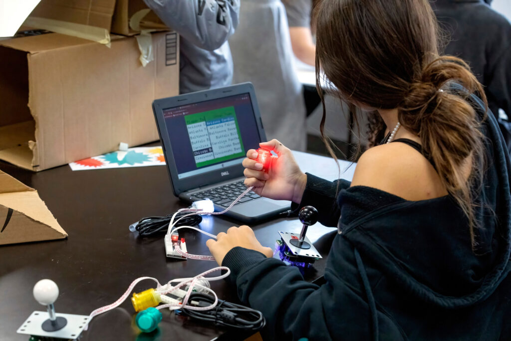 A student coding a game in front of a computer, with game controllers visible on the desk.