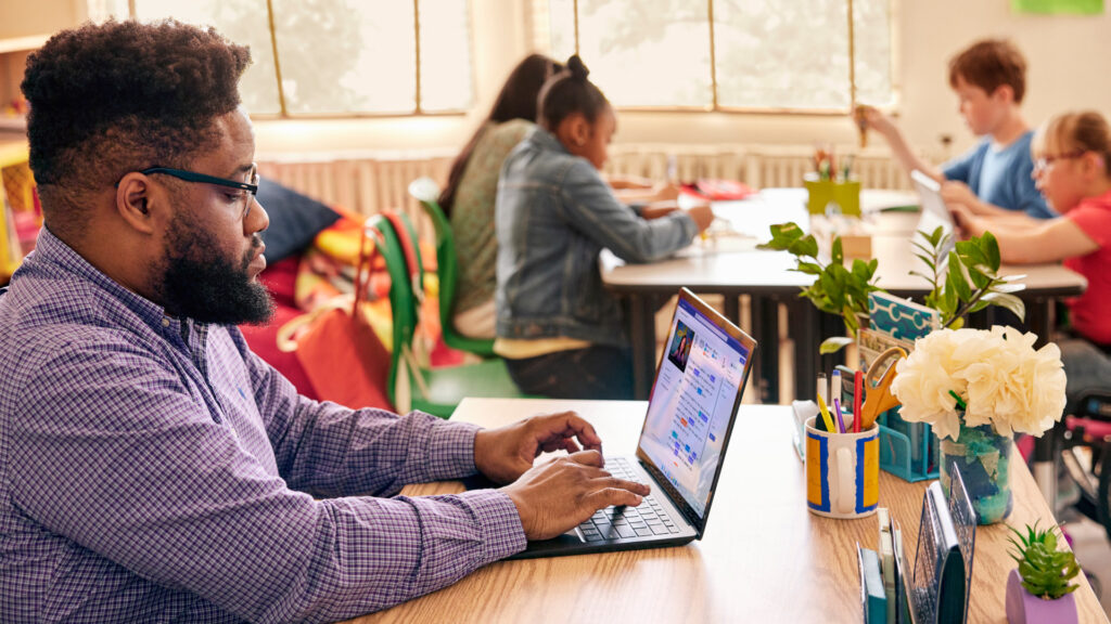 A teacher sitting at a desk in a classroom and working on a laptop with a group of students working at table in the background.