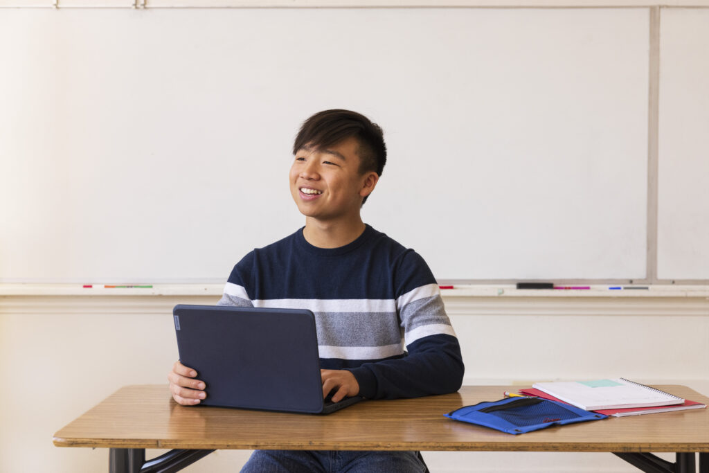 A teacher sitting at a desk at the front of a classroom with an open laptop in front of them.