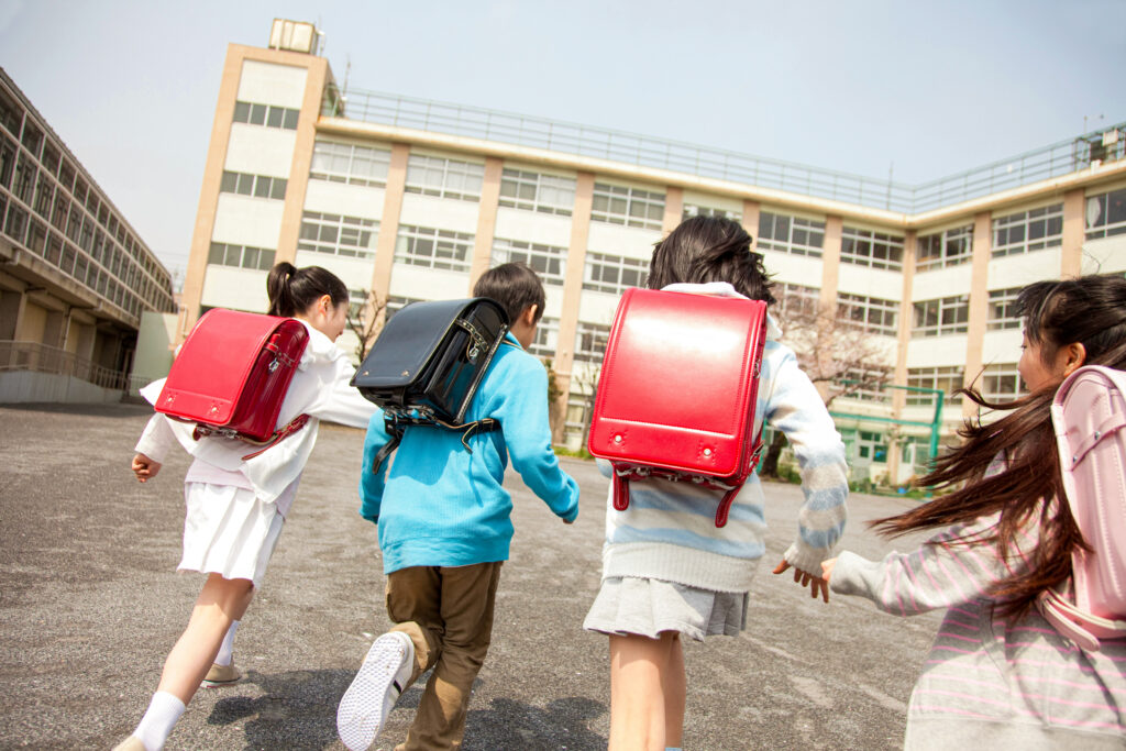 Four elementary school students wearing backpacks and walking across the schoolyard to the schoolhouse.