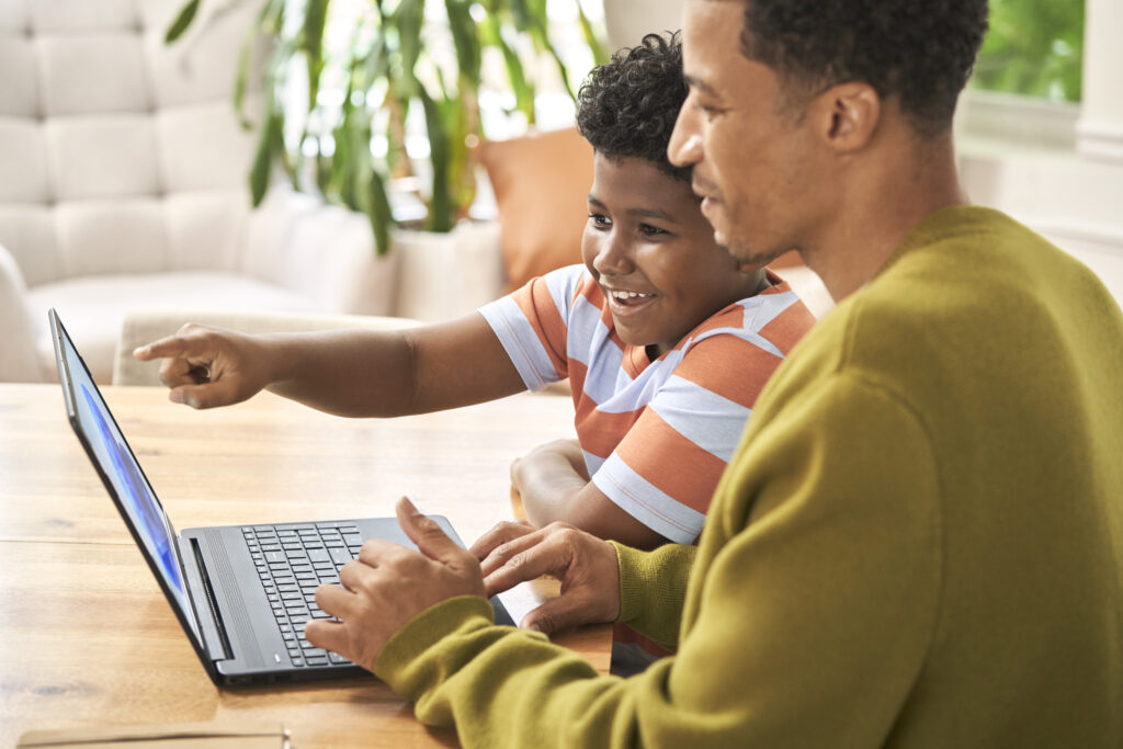 A parent helping a smiling child with homework as they both look at the laptop screen.