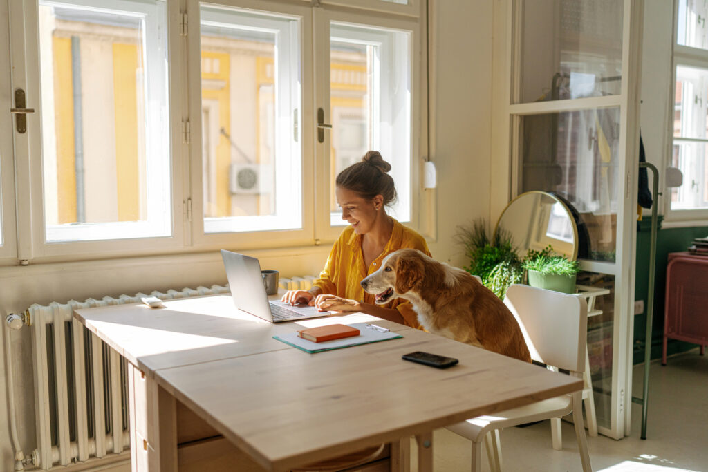 A woman sitting in a sunny apartment at a table with her dog sitting next to her, looking at her laptop.