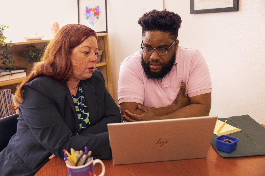 An educator and a school technologist sit at a desk in a school office, looking at a laptop screen together.