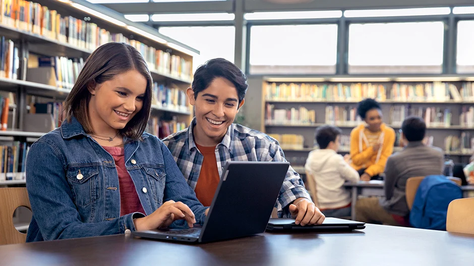 Two students working together on a laptop in a library.