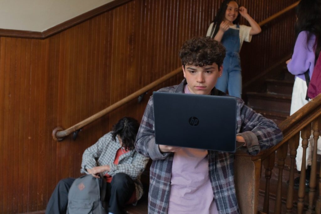 A male pre-teen student standing at the bottom of a staircase at school and working on a laptop. There are three other students hanging out on the stairs behind him.