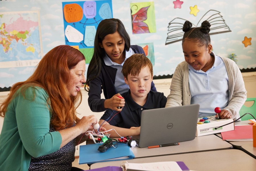 Decorative. A teacher engages with three students who are working on a laptop at a table in a school classroom.