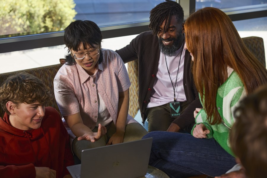 Decorative. A professor sits with a group of college students who are working together on a laptop.