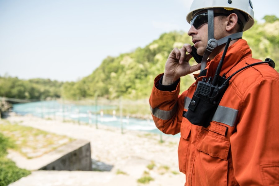 Male emergency response worker wearing a hardhat, sunglasses, walkie-talkie and jacket standing beside a body of water talking on a cellphone.
