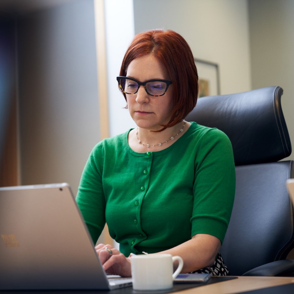A red-haired woman with a green shirt and glasses, sitting inside an office working on her laptop. She has a cup of coffee or tea and a phone by her side.