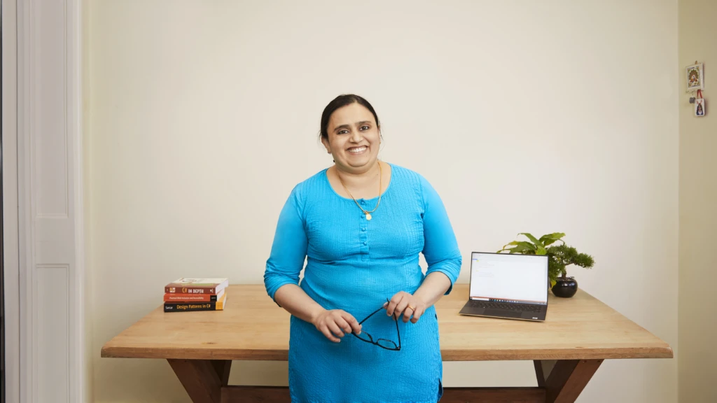 Poornima stands in front of her desk, smiling. On the desk are some books and an open laptop.