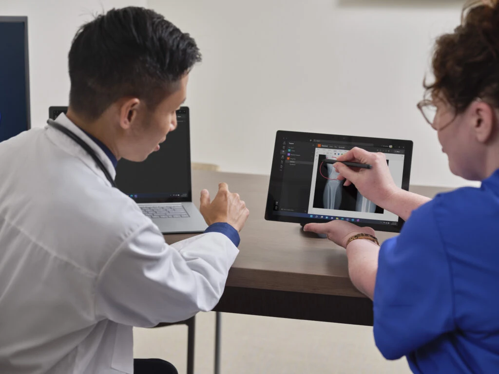 A nurse is showing a doctor an x-ray in Teams while holding a Surface Pro 8. A Laptop 4 is sitting on the desk.