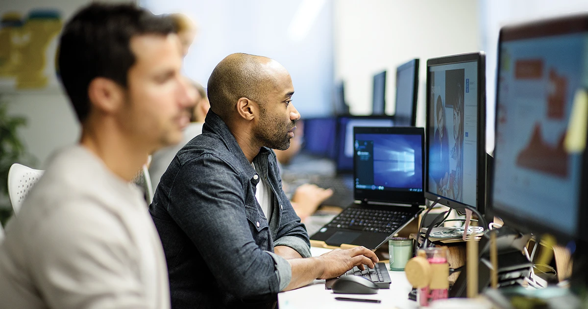 Two men sit at a long desk in front of several computer screens