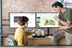 Two coworkers sit in front of a desk, looking at concept art on a desktop computer screen.