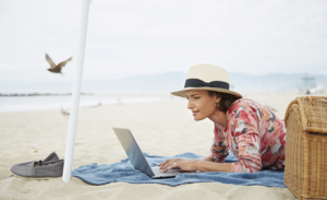 A woman works remotely under a parasol, laptop open on a beach towel.