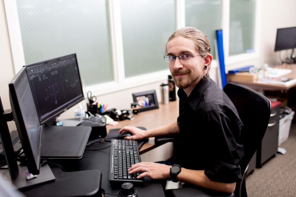 Portrait of male worker in manufacturing office sitting at desk in front of two desktop monitors. He is looking at camera, with one hand on the keyboard and one hand on the mouse. Screens show basic diagrams.