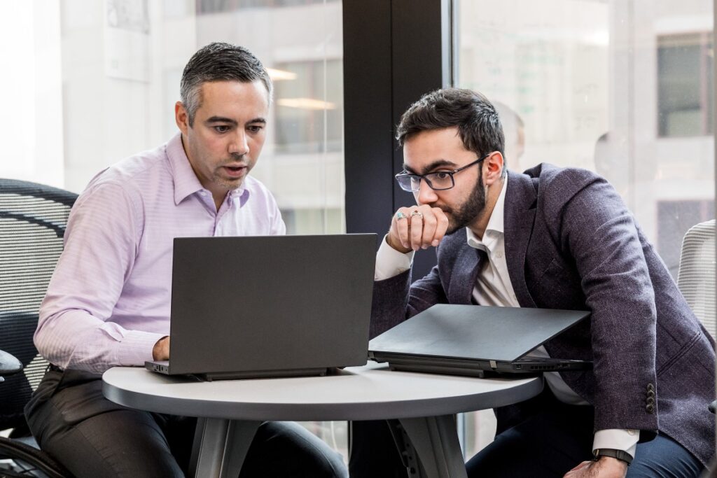 Two male office workers sitting at small round table in financial office, both looking at open laptop (screen not shown).