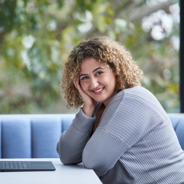 A person sitting at a table at their laptop, looking at the camera, smiling.