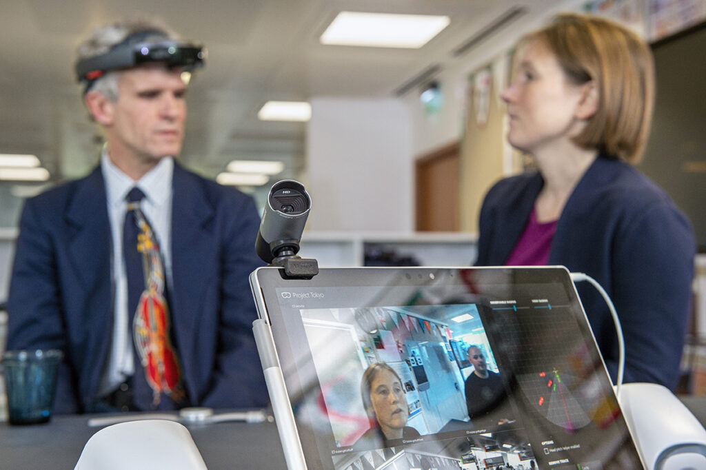 Peter Bosher, left background, an audio engineer who is blind, checks out the latest iteration of the Project Tokyo system at Microsoft’s research lab in Cambridge, UK. Bosher wears a modified Microsoft HoloLens that streams images of its visual field to computer hardware for processing. A dashboard on the laptop screen shows the visual field. Microsoft researchers Cecily Morrison, right background, and left on the screen, and Martin Grayson, right on the screen, are visible to the HoloLens. Photo by Jonathan Banks